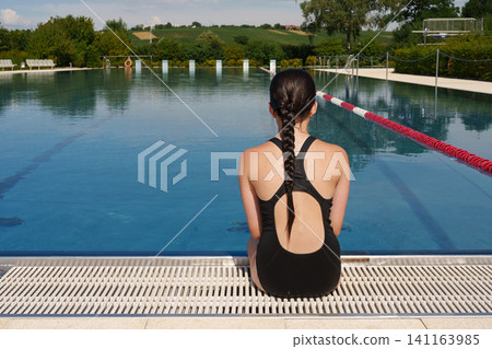 Teen girl sitting by outdoor pool and looking at water Teen girl sitting by outdoor pool and looking at water 141163985