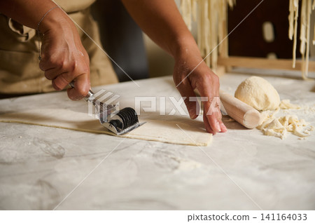 Woman cutting homemade pasta dough with a rolling pin and cutter in a rustic kitchen workspace Woman cutting homemade pasta dough with a rolling pin and cutter in a rustic kitchen workspace 141164033