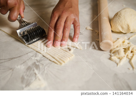 Hands cutting homemade pasta dough with rolling pin and pastry cutter on floured surface Hands cutting homemade pasta dough with rolling pin and pastry cutter on floured surface 141164034
