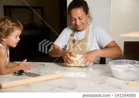 Mother and child kneading dough together at the kitchen table, smiling while baking Mother and child kneading dough together at the kitchen table, smiling while baking 141164080