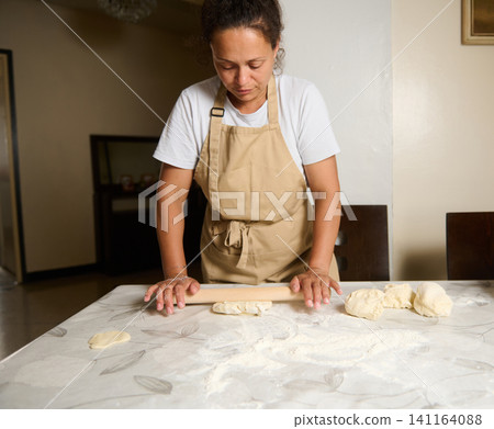 Woman rolling dough with a rolling pin on a flour-dusted table at home kitchen Woman rolling dough with a rolling pin on a flour-dusted table at home kitchen 141164088