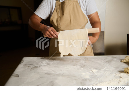 Woman rolling out dough with a wooden rolling pin in a home kitchen preparing flatbread Woman rolling out dough with a wooden rolling pin in a home kitchen preparing flatbread 141164091