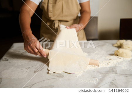 Woman rolling dough with a rolling pin on a floured kitchen table while preparing homemade pastry Woman rolling dough with a rolling pin on a floured kitchen table while preparing homemade pastry 141164092