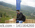 Young Indian male traveler sitting on a bamboo deck overlooking the scenic tea gardens of Tinchuley Darjeeling 141164260