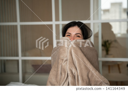 Woman wrapped in a cozy blanket sitting in a modern home interior during winter. Cold weather home comfort and energy saving solutions Woman wrapped in a cozy blanket sitting in a modern home interior during winter. Cold weather home comfort and energy saving solutions 141164349