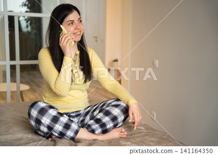 Young woman in casual attire sitting on bed having a phone conversation at home. Mobile lifestyle remote communication and home consultation Young woman in casual attire sitting on bed having a phone conversation at home. Mobile lifestyle remote communication and home consultation 141164350