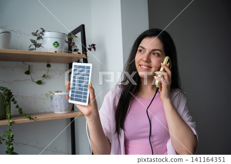 Smiling woman holding a solar panel and talking on the phone in a home office. Solar energy consultation renewable power and customer support Smiling woman holding a solar panel and talking on the phone in a home office. Solar energy consultation renewable power and customer support 141164351