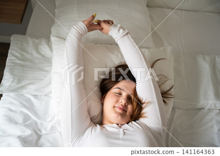 Young woman stretching in bed with a relaxed expression in the morning light. Morning wellness relaxation and waking up routine Young woman stretching in bed with a relaxed expression in the morning light. Morning wellness relaxation and waking up routine 141164363