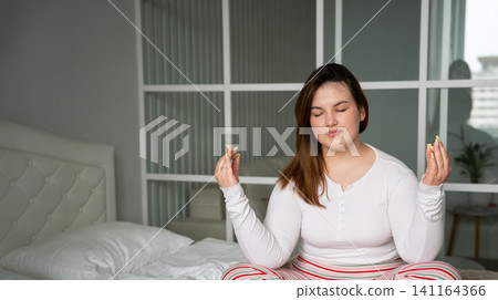 Young woman meditating peacefully on bed in a bright bedroom. Mindfulness self development and relaxation in a home environment 141164366