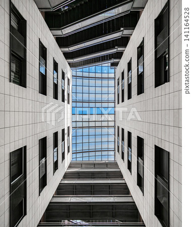 Upward architectural view through narrow atrium beneath glass skylight framed by white concrete walls, sharp geometric angles, clean lines, strong symmetry, cool daylight, modern urban structure Upward architectural view through narrow atrium beneath glass skylight framed by white concrete walls, sharp geometric angles, clean lines, strong symmetry, cool daylight, modern urban structure 141164528