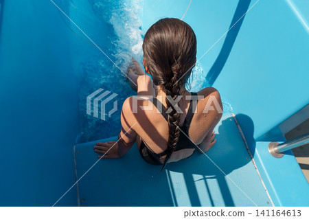 Teen girl sliding down blue water slide at outdoor pool in summer Teen girl sliding down blue water slide at outdoor pool in summer 141164613