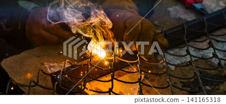 Worker wearing protective gloves performing arc welding on a metal fence, generating intense light, sparks, and smoke, showcasing industrial repair and construction activity. Worker wearing protective gloves performing arc welding on a metal fence, generating intense light, sparks, and smoke, showcasing industrial repair and construction activity. 141165318