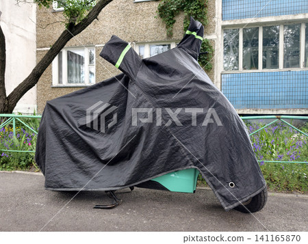 Black nylon motorcycle cover covering a motorbike parked in a quiet residential yard near an apartment building designed for weather resistance and outdoor storage safety Black nylon motorcycle cover covering a motorbike parked in a quiet residential yard near an apartment building designed for weather resistance and outdoor storage safety 141165870