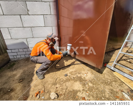 Worker wearing a respirator mask and protective clothing, using a spray gun to apply paint on a large metal surface outside a building during a renovation project. Worker wearing a respirator mask and protective clothing, using a spray gun to apply paint on a large metal surface outside a building during a renovation project. 141165931