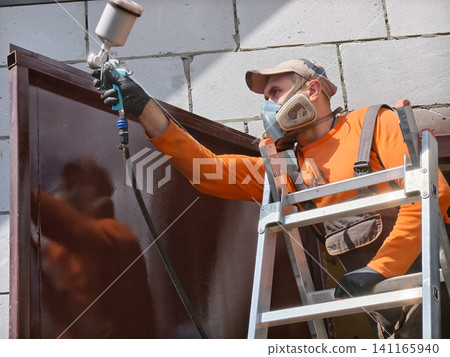 Worker wearing a respirator, cap, and protective gloves is standing on a ladder and applying brown spray paint to a metal garage door, performing a professional renovation on a building exterior. Worker wearing a respirator, cap, and protective gloves is standing on a ladder and applying brown spray paint to a metal garage door, performing a professional renovation on a building exterior. 141165940
