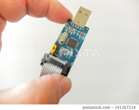 Hand holding a blue USB microcircuit electronic board programmer with ribbon cable. Hand holding a blue USB microcircuit electronic board programmer with ribbon cable. 141167114