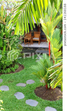 Tropical garden pathway framed by palm fronds, leading toward wooden patio chairs and small table in sunny courtyard with lush greenery, dappled light, and calm resort style Tropical garden pathway framed by palm fronds, leading toward wooden patio chairs and small table in sunny courtyard with lush greenery, dappled light, and calm resort style 141168188