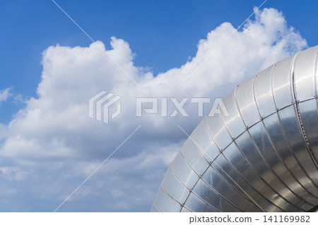 Close-up of a large silver metallic insulation pipe elbow at a petrochemical plant against a bright blue sky with white clouds. Close-up of a large silver metallic insulation pipe elbow at a petrochemical plant against a bright blue sky with white clouds. 141169982