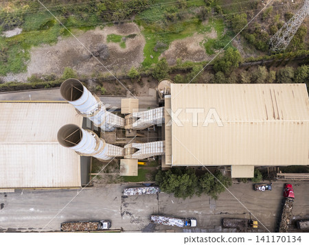 Aerial drone view of an industrial facility with large smokestacks, factory rooftops, service roads, and waste transport trucks 141170134