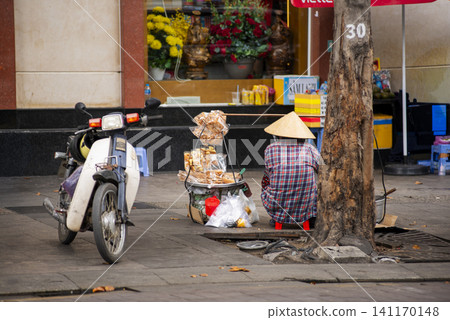Street vendor wearing a traditional conical hat selling snacks beside a motorbike on a sidewalk in Ho Chi Minh City, Vietnam 141170148