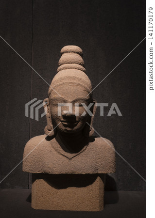 Serene smile of an ancient stone Buddha statue bust displayed in a museum with dramatic lighting. Serene smile of an ancient stone Buddha statue bust displayed in a museum with dramatic lighting. 141170179