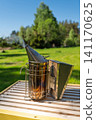 Bee smoker tool emitting smoke on top of a wooden beehive frame in a sunny rural apiary field 141170625