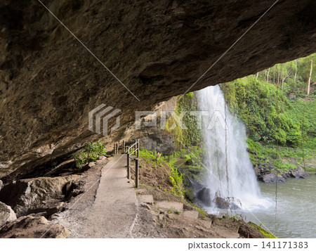 Waterfall flows beside path under rock in forest during daytime Waterfall flows beside path under rock in forest during daytime 141171383