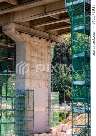 A photograph taken from a low angle, showing the bridge piers of an elevated road and scaffolding with green safety nets. 141171933
