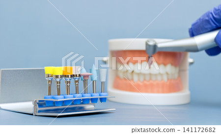 Set of colorful dental polishing burs in sharp focus. Blurred dentist hand holding drill handpiece treating jaw model in background. Set of colorful dental polishing burs in sharp focus. Blurred dentist hand holding drill handpiece treating jaw model in background. 141172682
