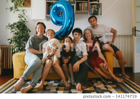 Happy multigenerational family gathered on a sofa during a boy's birthday celebration at home, with a large blue number nine balloon and pet dog. Happy multigenerational family gathered on a sofa during a boy's birthday celebration at home, with a large blue number nine balloon and pet dog. 141174719