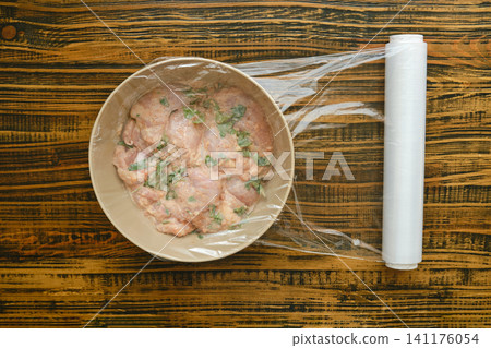 Marinating chicken with herbs and spice on wooden table in kitchen by covering the bowl with cling film Marinating chicken with herbs and spice on wooden table in kitchen by covering the bowl with cling film 141176054