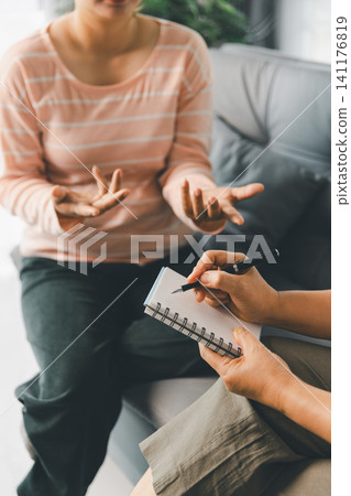 Female client explains feelings with open hands while therapist writes in notebook during counseling, showing talk therapy, active listening, emotional support, trust, guidance, and mental wellness. Female client explains feelings with open hands while therapist writes in notebook during counseling, showing talk therapy, active listening, emotional support, trust, guidance, and mental wellness. 141176819