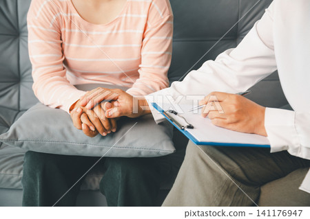 Therapist gently holds female client's hands while writing notes during counseling, showing empathy, reassurance, mental health support, professional care, and trusted consultation indoors. Therapist gently holds female client's hands while writing notes during counseling, showing empathy, reassurance, mental health support, professional care, and trusted consultation indoors. 141176947