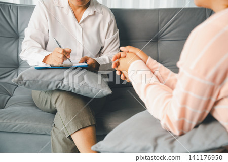 Therapist writes notes on clipboard while female client sits with clasped hands during private counseling, showing professional mental health care, empathy, trust, and guidance indoors. Therapist writes notes on clipboard while female client sits with clasped hands during private counseling, showing professional mental health care, empathy, trust, and guidance indoors. 141176950