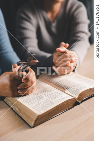 Close up of a person holding a wooden cross while praying over an open Bible, Christian faith, worship, hope, salvation, and devotion concept. Close up of a person holding a wooden cross while praying over an open Bible, Christian faith, worship, hope, salvation, and devotion concept. 141176951