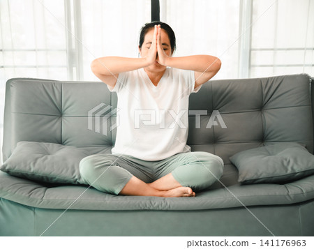 Middle aged Asian woman practicing meditation and breathing exercise on sofa at home, concept of yoga, mindfulness, wellness, stress relief, healthy lifestyle, and self care. Middle aged Asian woman practicing meditation and breathing exercise on sofa at home, concept of yoga, mindfulness, wellness, stress relief, healthy lifestyle, and self care. 141176963