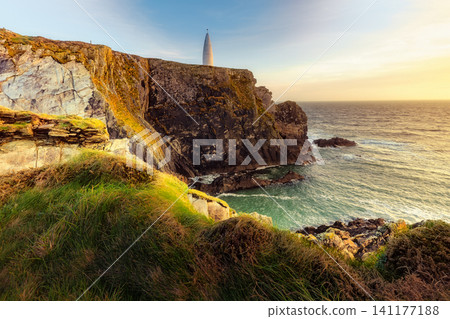 The sun shines brightly over a rugged coastline featuring steep cliffs covered in grass and the sea below. 141177188