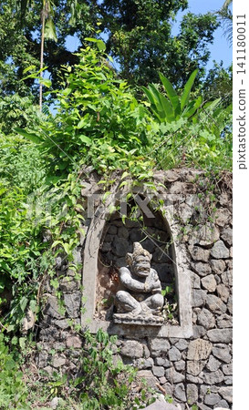 Carved stone guardian statue seated in arched niche set into weathered rock wall, framed by tropical greenery, climbing vines, moss and bright sunlight in lush outdoor setting 141180011