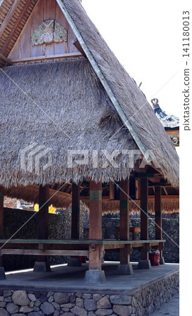 Traditional Balinese pavilion with steep thatched roof, carved wooden ornament, timber posts, raised stone base, and open shaded interior in tropical village setting Traditional Balinese pavilion with steep thatched roof, carved wooden ornament, timber posts, raised stone base, and open shaded interior in tropical village setting 141180013