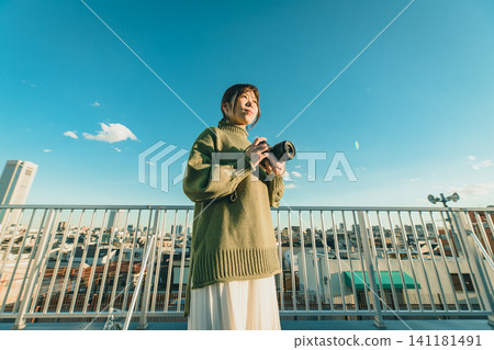 A woman with a camera gazes at the cityscape under a blue sky. A woman with a camera gazes at the cityscape under a blue sky. 141181491