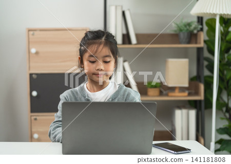 Young girl focused and working on laptop computer at desk in modern home interior with bookshelf and lamp background Young girl focused and working on laptop computer at desk in modern home interior with bookshelf and lamp background 141181808