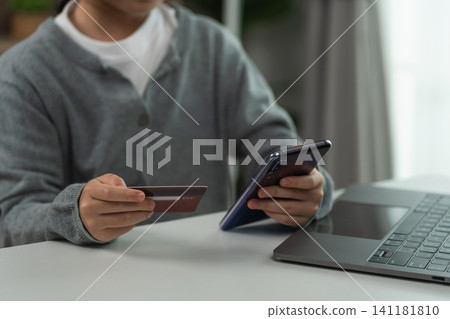 Young person holding credit card making online payment with smartphone beside open laptop on white desk at home Young person holding credit card making online payment with smartphone beside open laptop on white desk at home 141181810