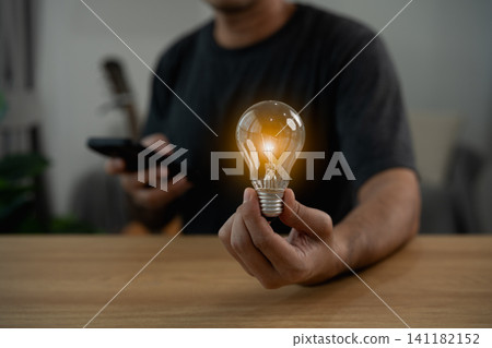 Person holding glowing light bulb symbolizing creative idea and innovation with blurred background and smartphone in hand Person holding glowing light bulb symbolizing creative idea and innovation with blurred background and smartphone in hand 141182152