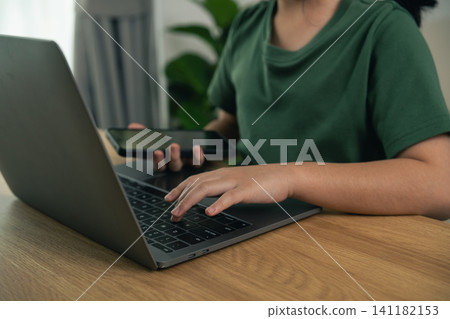 Young person using laptop and holding smartphone while sitting at wooden desk with blurred background indoors 141182153