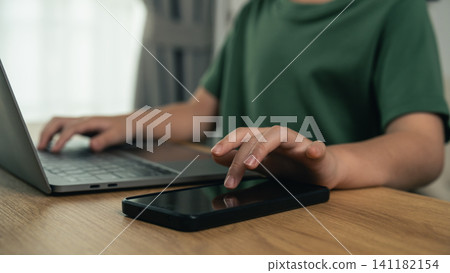 Person using smartphone and laptop simultaneously on wooden table for multitasking digital communication in casual green shirt 141182154