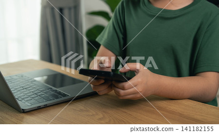 Child in green shirt holding black smartphone using device near open laptop on wooden table indoors 141182155