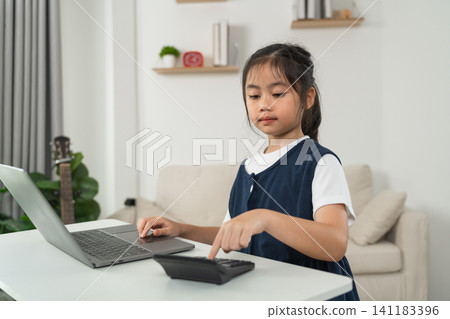 Young girl sitting at white table using laptop and pressing buttons on calculator focused on learning and doing homework in cozy living room with natural light Young girl sitting at white table using laptop and pressing buttons on calculator focused on learning and doing homework in cozy living room with natural light 141183396