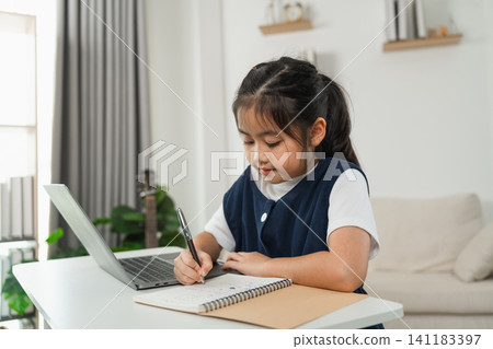 Young girl focused on writing in notebook while using laptop on white table at home learning environment with natural light and modern interior background Young girl focused on writing in notebook while using laptop on white table at home learning environment with natural light and modern interior background 141183397
