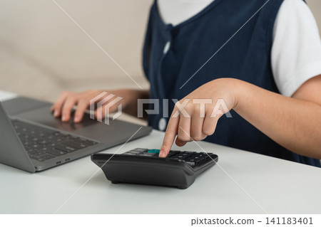 Child using calculator and laptop on white desk for learning numerical skills with focused hand pressing button in education and technology setting 141183401