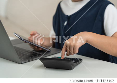 Child using calculator and holding smartphone while sitting in front of laptop working on math homework or financial calculation at white desk indoors Child using calculator and holding smartphone while sitting in front of laptop working on math homework or financial calculation at white desk indoors 141183402